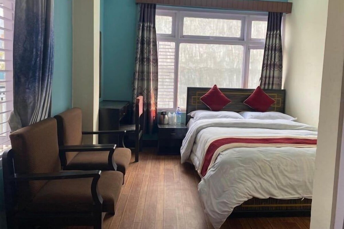 Una habitación de hotel con cama, sillón, mesita de noche y ventana con cortinas.