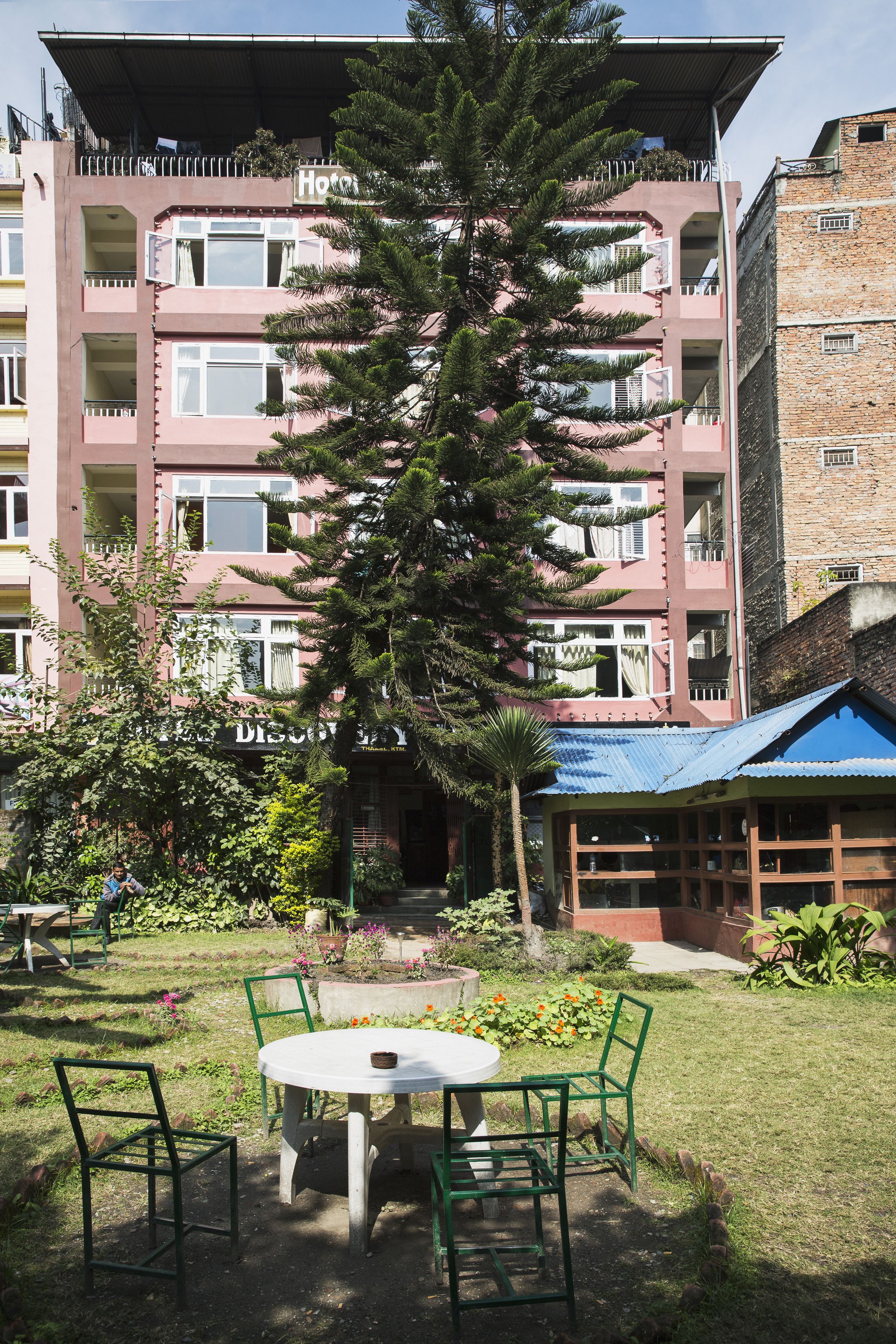 A hotel with a balcony, a garden with a table and chairs, and a large tree.