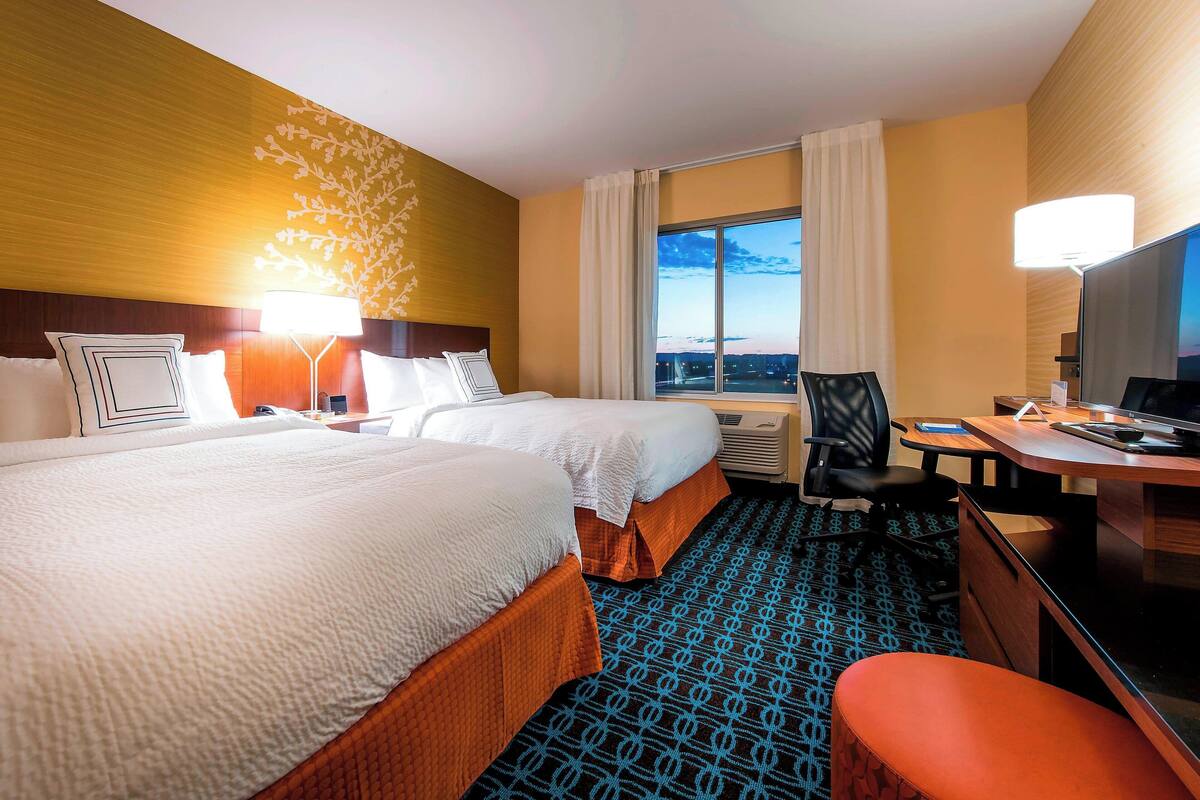 A hotel room with two beds, a desk, a chair, and a TV.