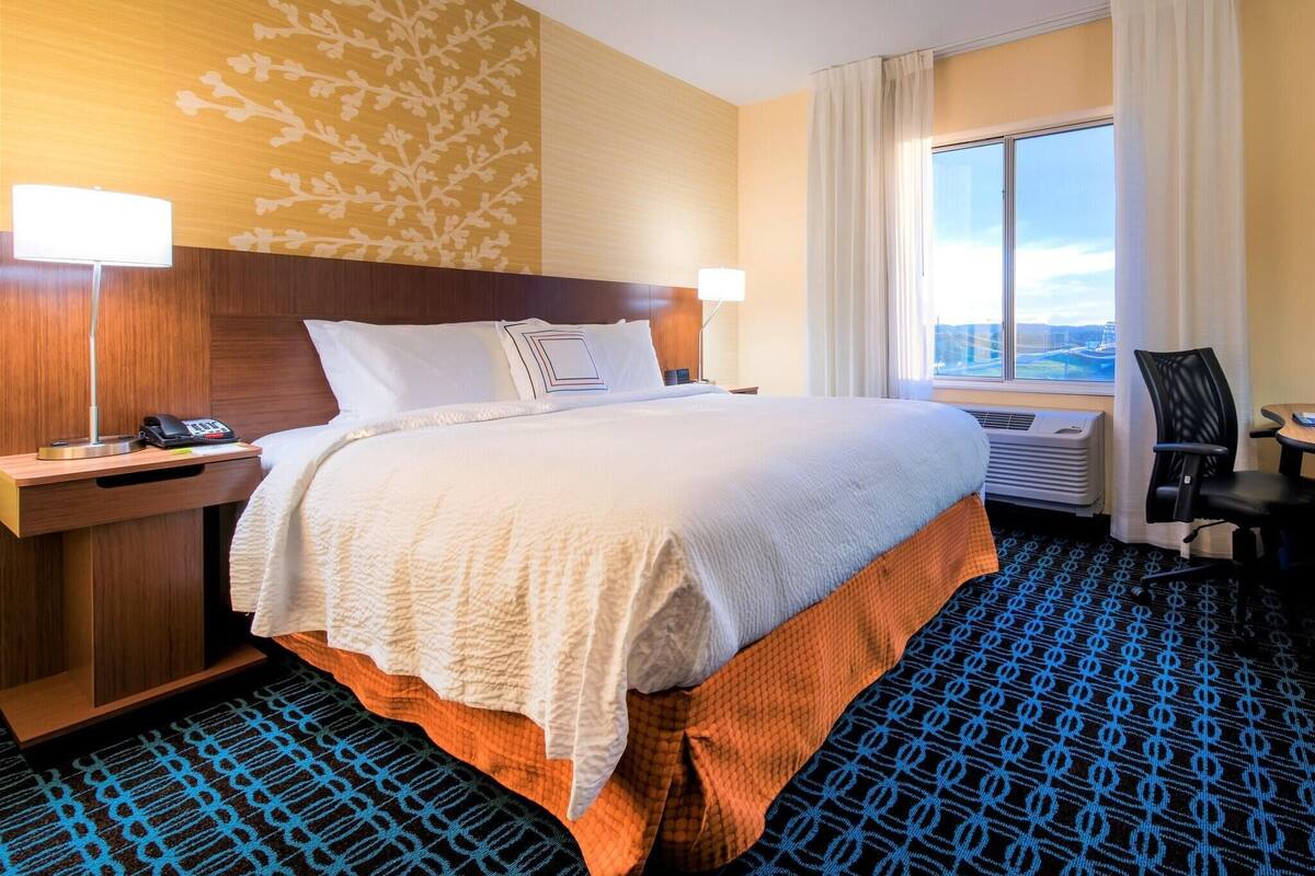 A hotel room with a bed, bedside lamps, a desk with a phone, a chair, and a large window with a view.
