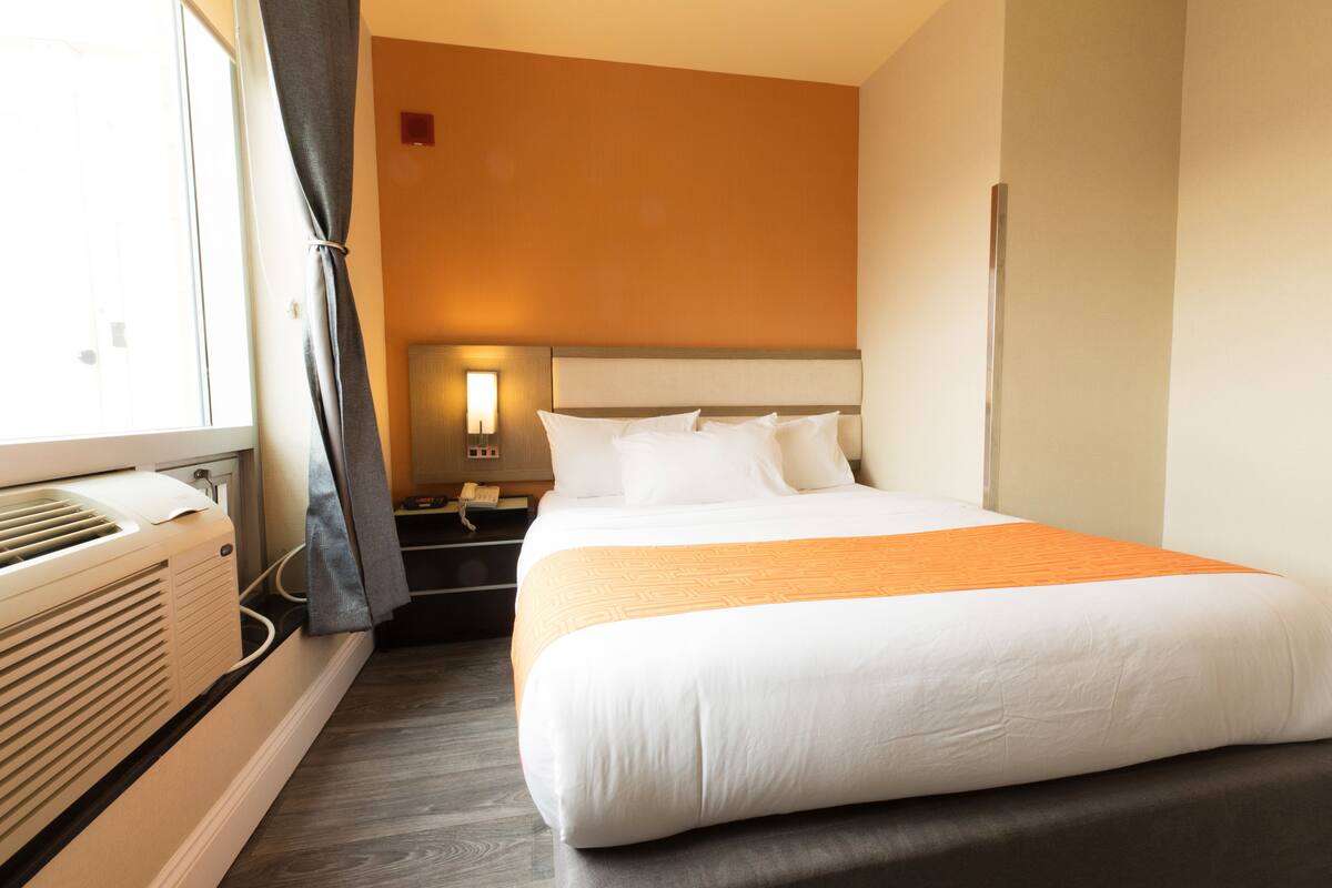 Hotellihuone, jossa on suuri sänky, yöpöytä lampulla, lämmitin ja ikkuna, jossa on verhot.