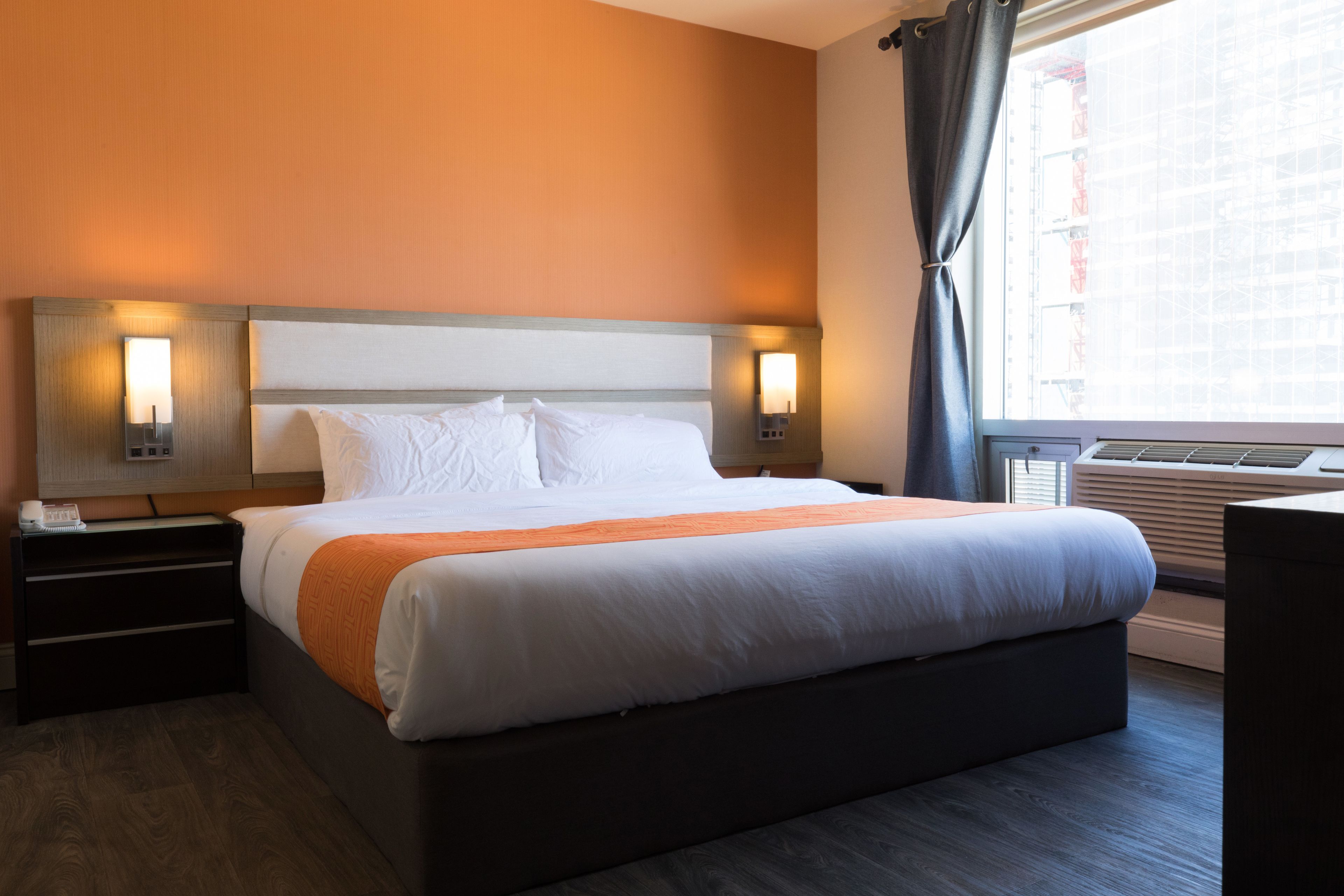 A hotel room with a large bed, two bedside tables, a nightstand, a window with curtains, and an air conditioning unit.
