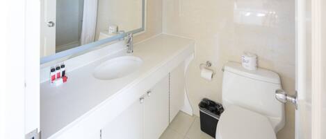 A bathroom with a white sink, mirror, and toilet. There are towels and a trash bin visible.