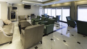 A modern hotel lobby with leather sofas, a water dispenser, a flat-screen TV, and large windows with blinds. - Romana Hotel - NYC (Long Island City)