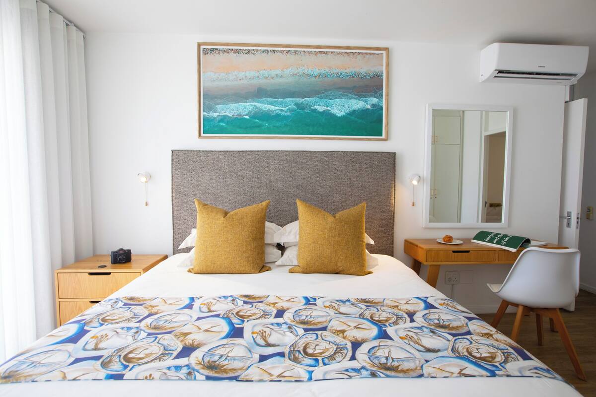 Quarto de hotel com cama, escrivaninha, cadeira, espelho e pintura de uma paisagem marítima.