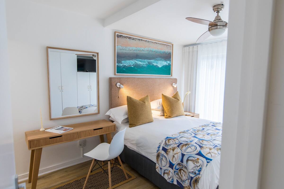 Quarto com cama, escrivaninha, cadeira, espelho e ventilador de teto.