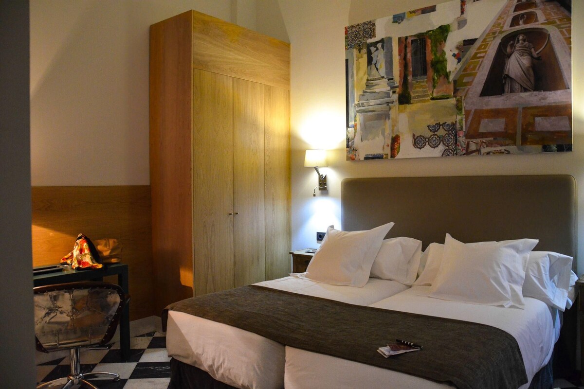 A hotel room with a bed, a wooden wardrobe, a desk with a chair, and a wall-mounted lamp.