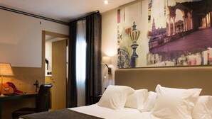 A hotel room with a bed, a desk, and a chair. There is a painting on the wall and a lamp on the desk. - Aguilas5 SevillaSuites (Seville)