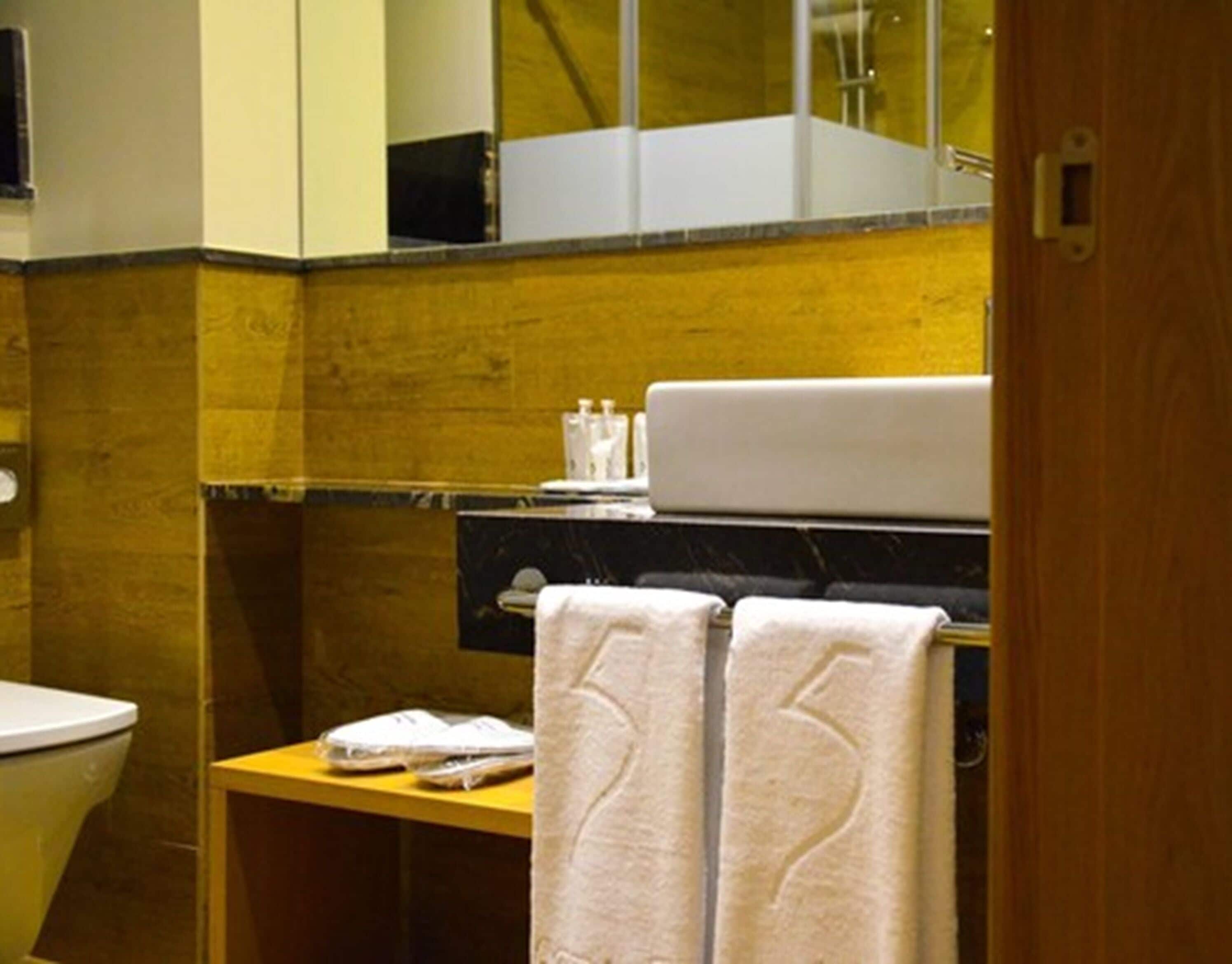 A bathroom with a toilet, a sink, and a mirror. There are towels hanging on the wall and a small table with toiletries.