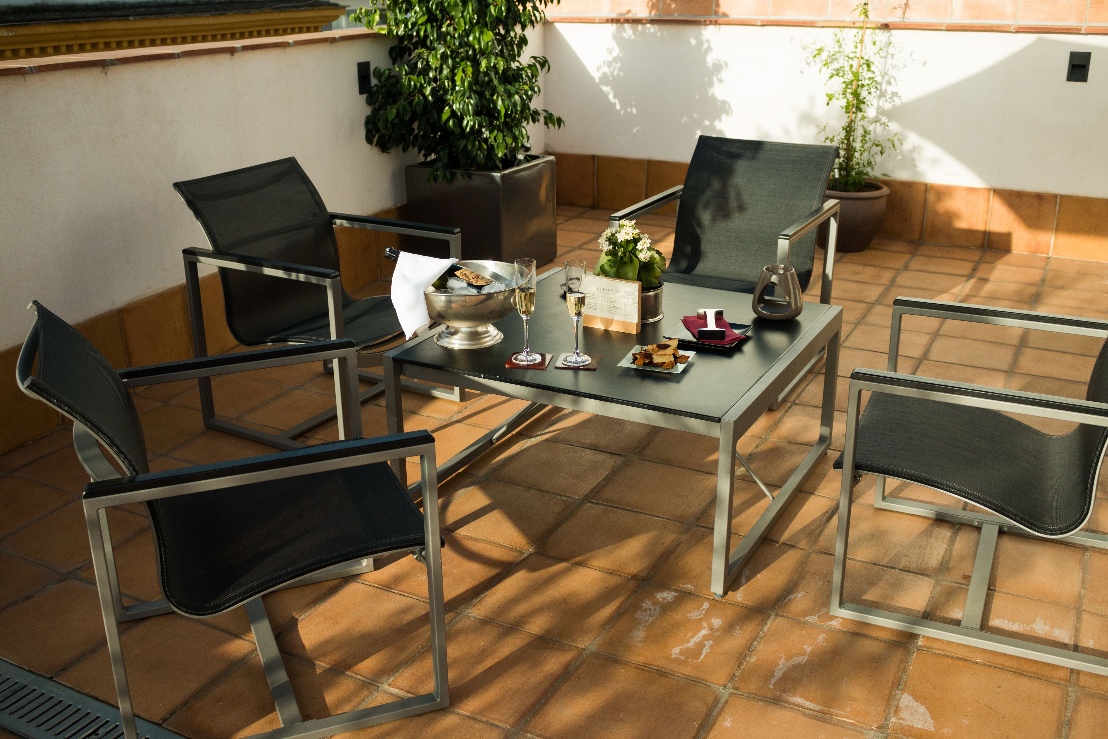 Eine Terrasse mit Tisch für zwei Personen, umgeben von vier Stühlen, einem Blumentopf sowie einem kleinen Tisch mit Kerze und Speisekarte.