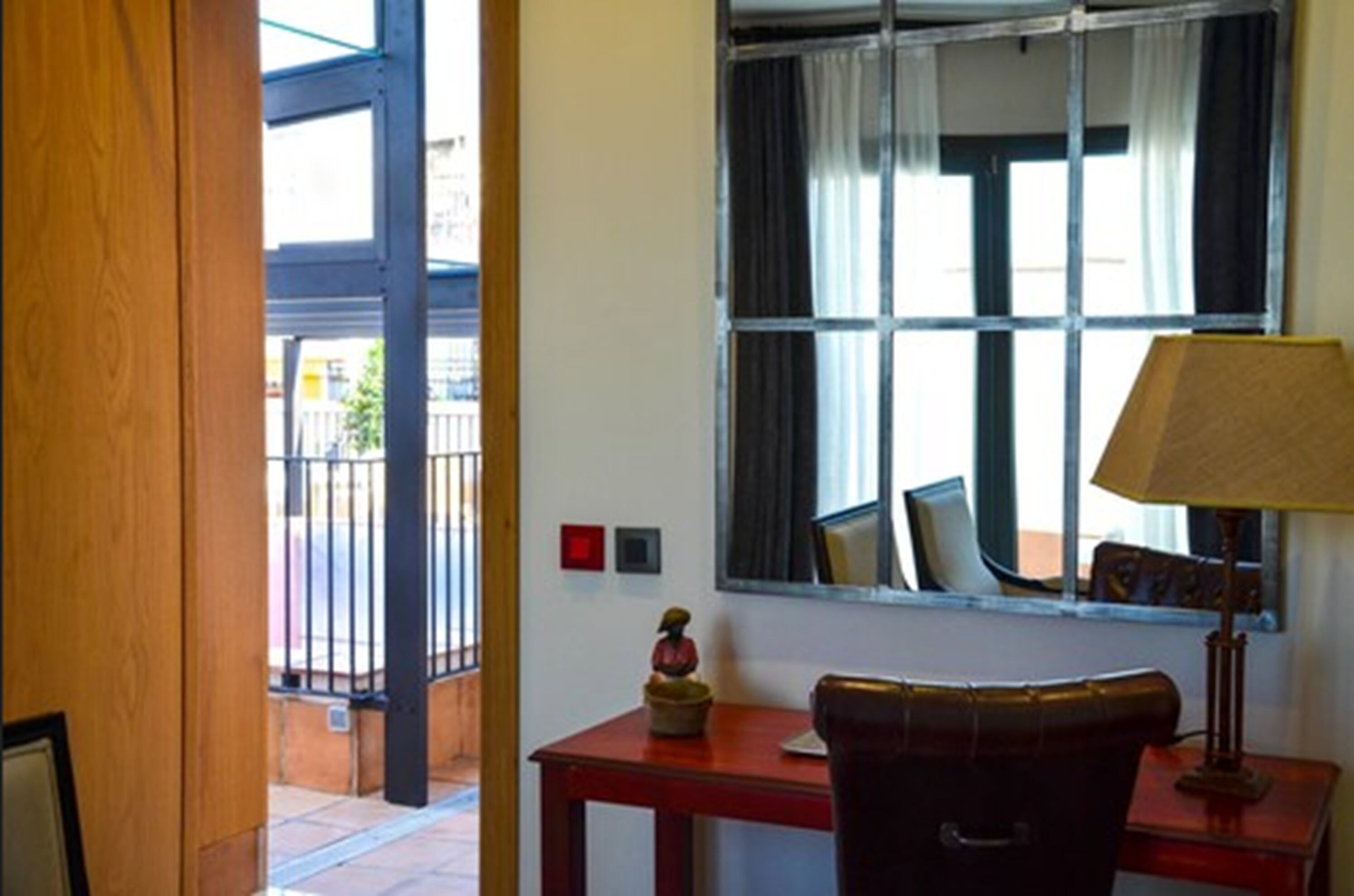 A hotel room with a wooden door, a window with curtains, a table with a lamp, and a small statue.