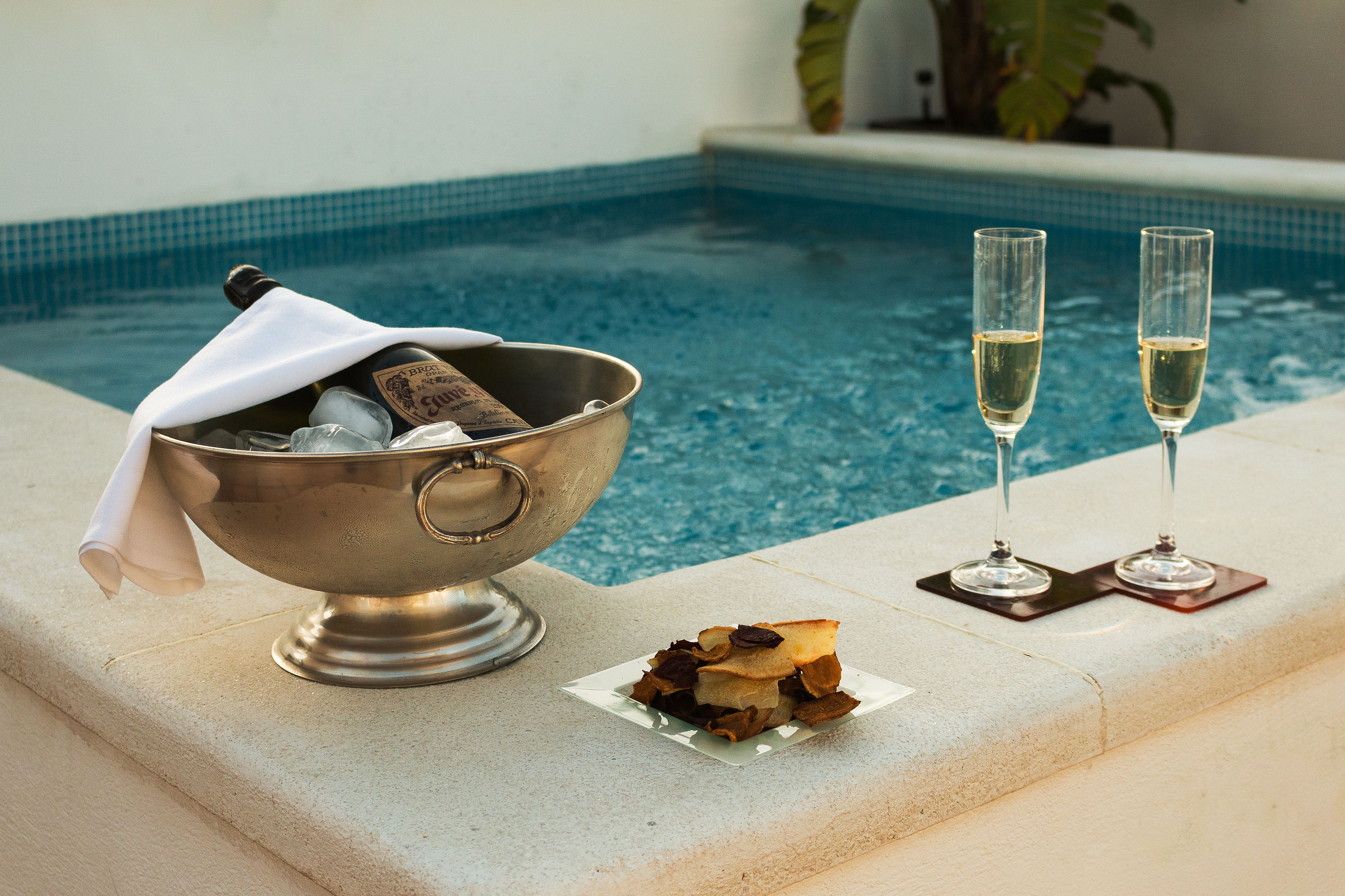 A poolside setting with a metal bowl containing bottles and glasses, and two glasses of champagne on a tray.