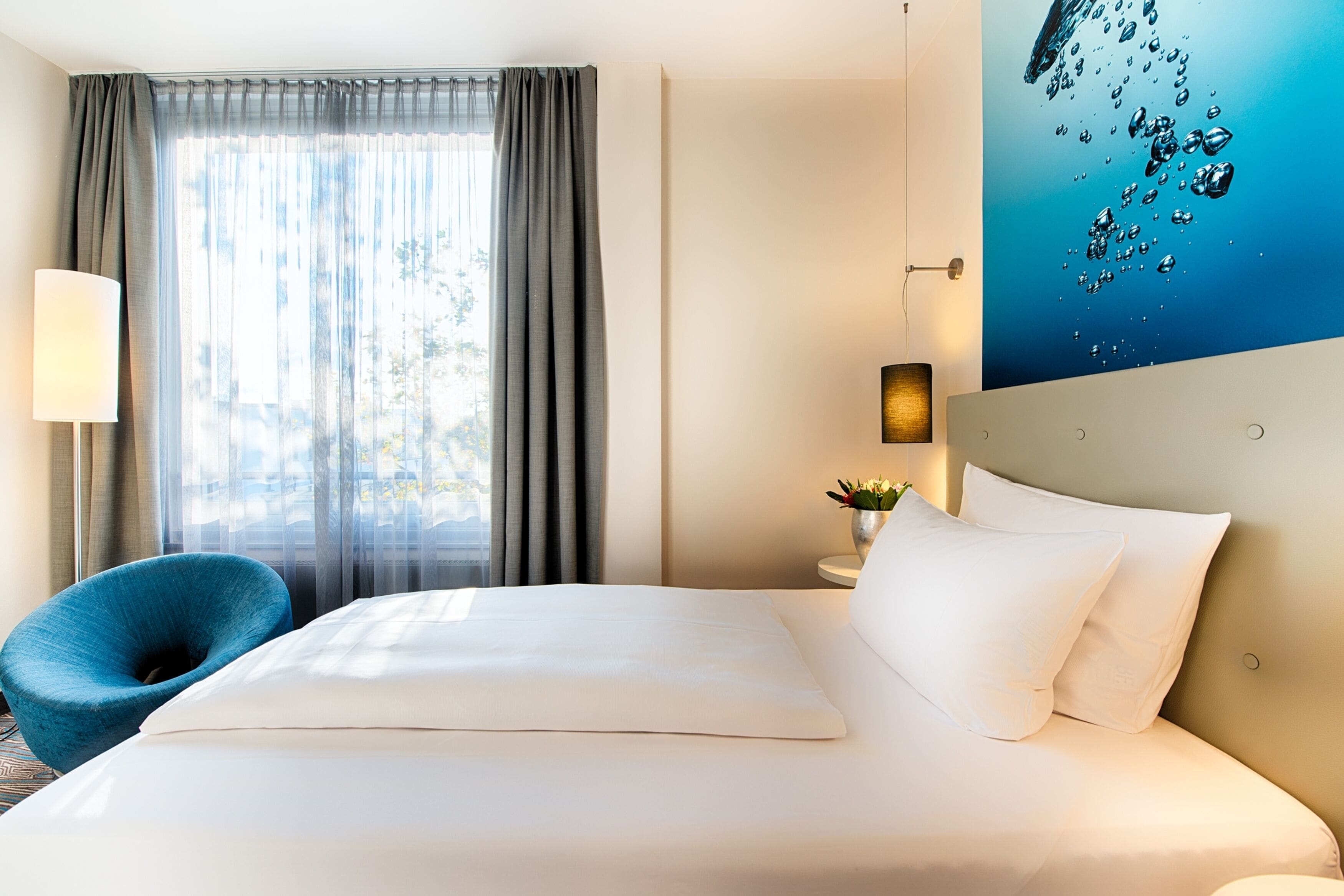 A hotel room with a large bed, a blue chair, a window with curtains, a wall-mounted lamp, and a painting of water bubbles.