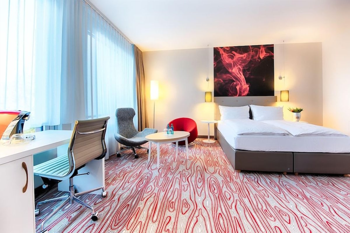 A hotel room with a bed, desk, chair, and a large abstract painting on the wall.