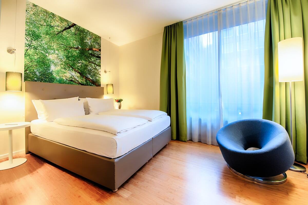 Une chambre d’hôtel moderne, dotée d’un lit, d’un fauteuil rond, d’une lampe fixée au mur et d’une grande fenêtre ornée de rideaux légers.