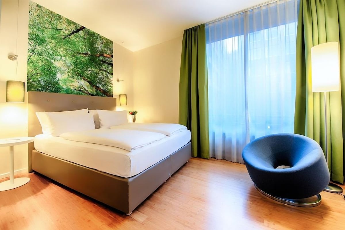 Una habitación de hotel con una cama, una silla negra redonda, un cuadro en la pared, una ventana con cortinas verdes y una lámpara de noche.
