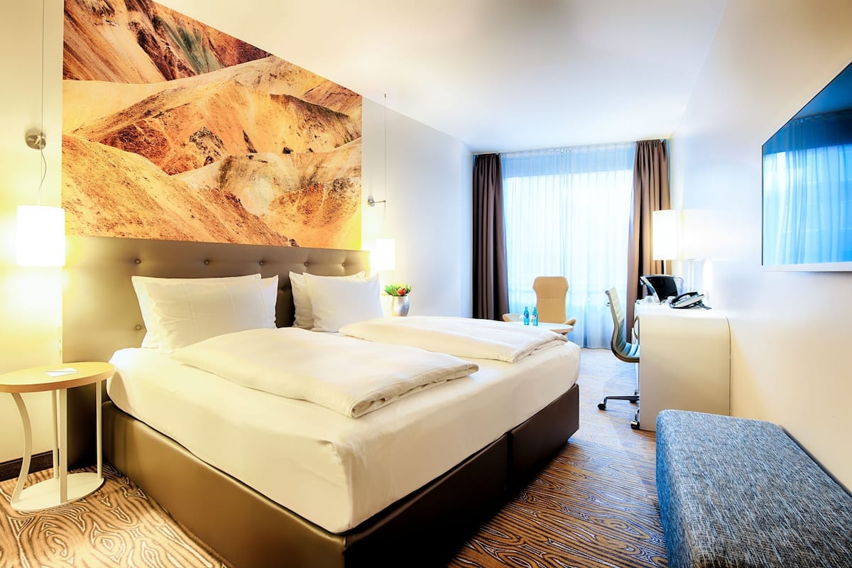 Une chambre d’hôtel avec un grand lit, un bureau, une chaise et un grand tableau abstrait accroché au mur.