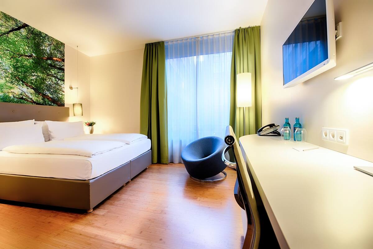 Une chambre d’hôtel comprenant un lit, une chaise, un bureau, une fenêtre avec des rideaux et un téléviseur fixé au mur.