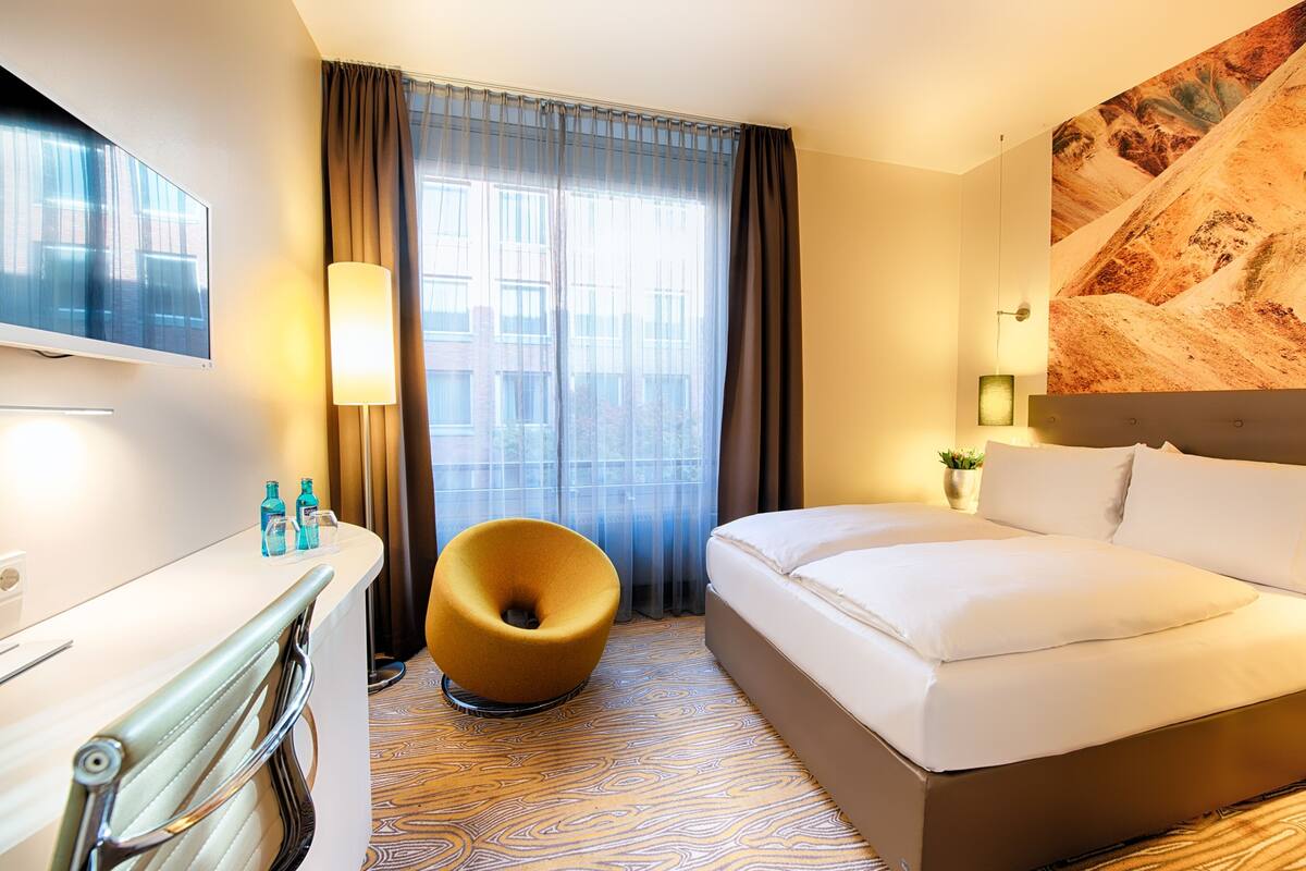 Une chambre d’hôtel avec un lit, une chaise, une télévision, un bureau et une fenêtre avec des rideaux.