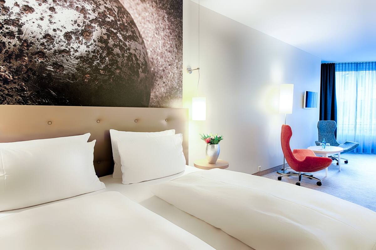 Une chambre d’hôtel avec un grand lit, une chaise rouge, une petite table et un grand tableau accroché au mur.