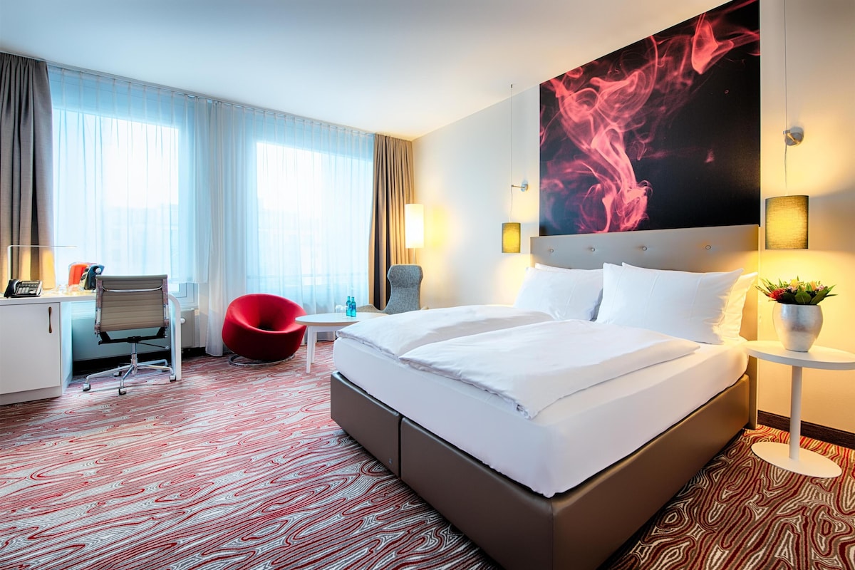A hotel room with a large bed, a desk with a chair, a red chair, and a large abstract painting on the wall.