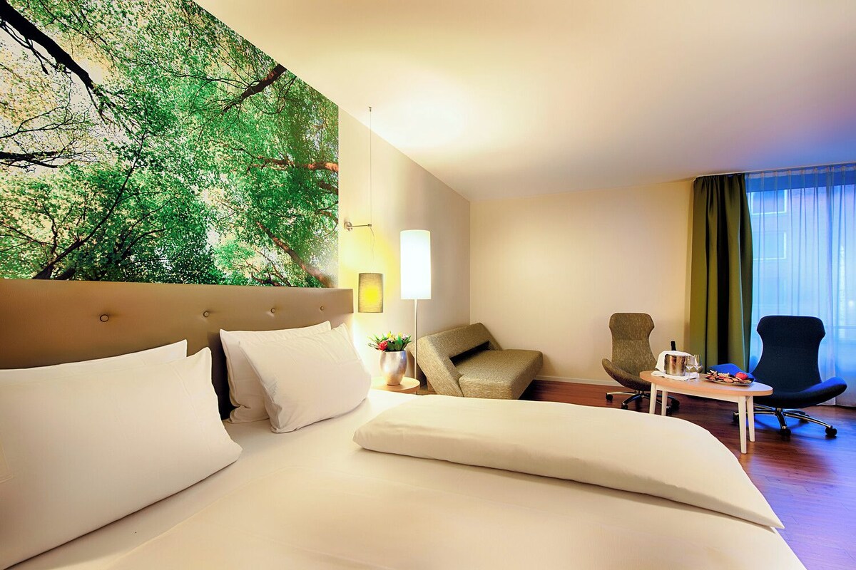 Habitación de hotel con cama, dos sillones, una mesita y una pared decorada con un mural de motivos naturales.