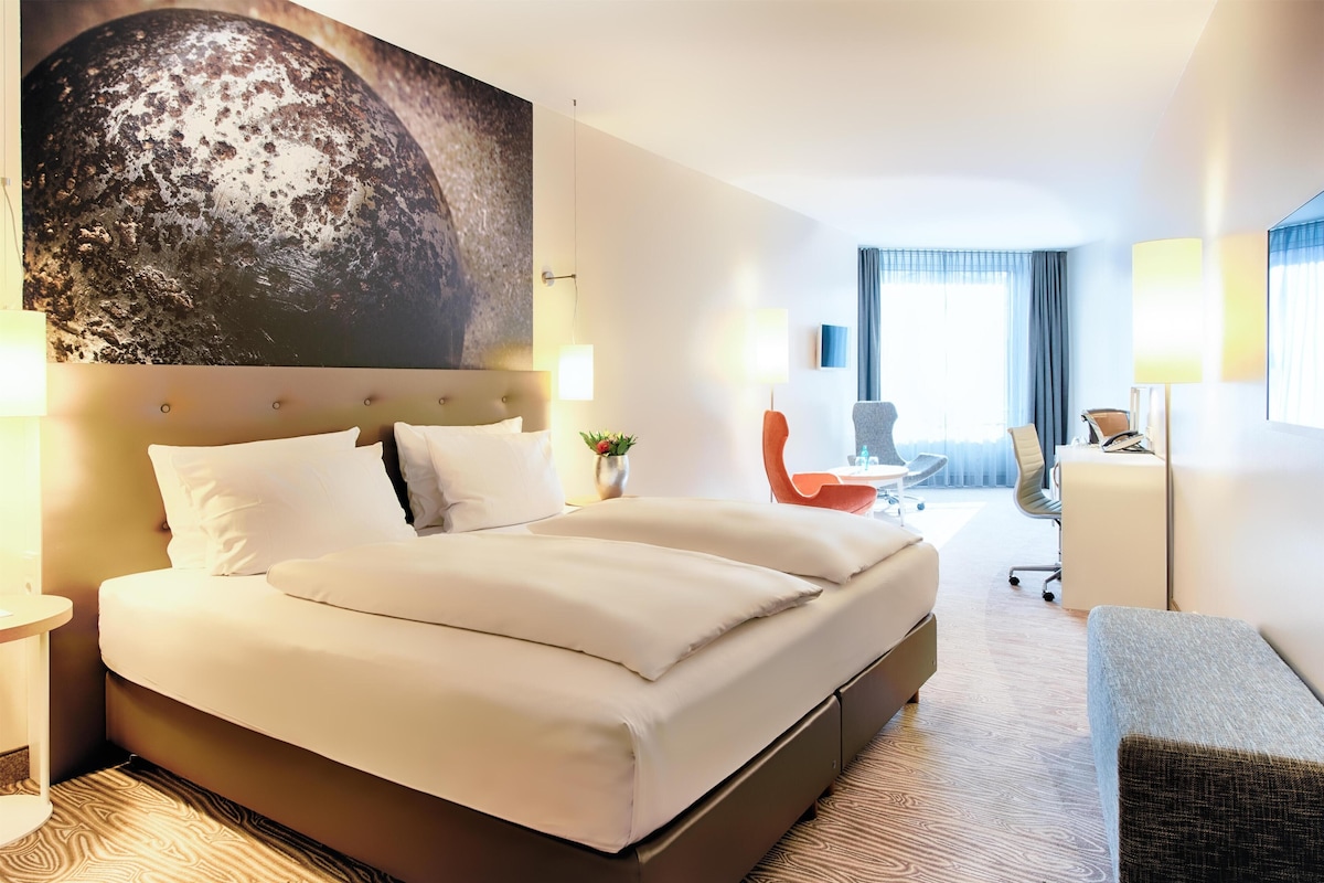 A hotel room with a large bed, a desk, a chair, and a view of the city.