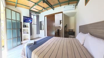 Un dormitorio con cama, televisor montado en la pared, baño con lavamanos y espejo, y una puerta que da a otra habitación.