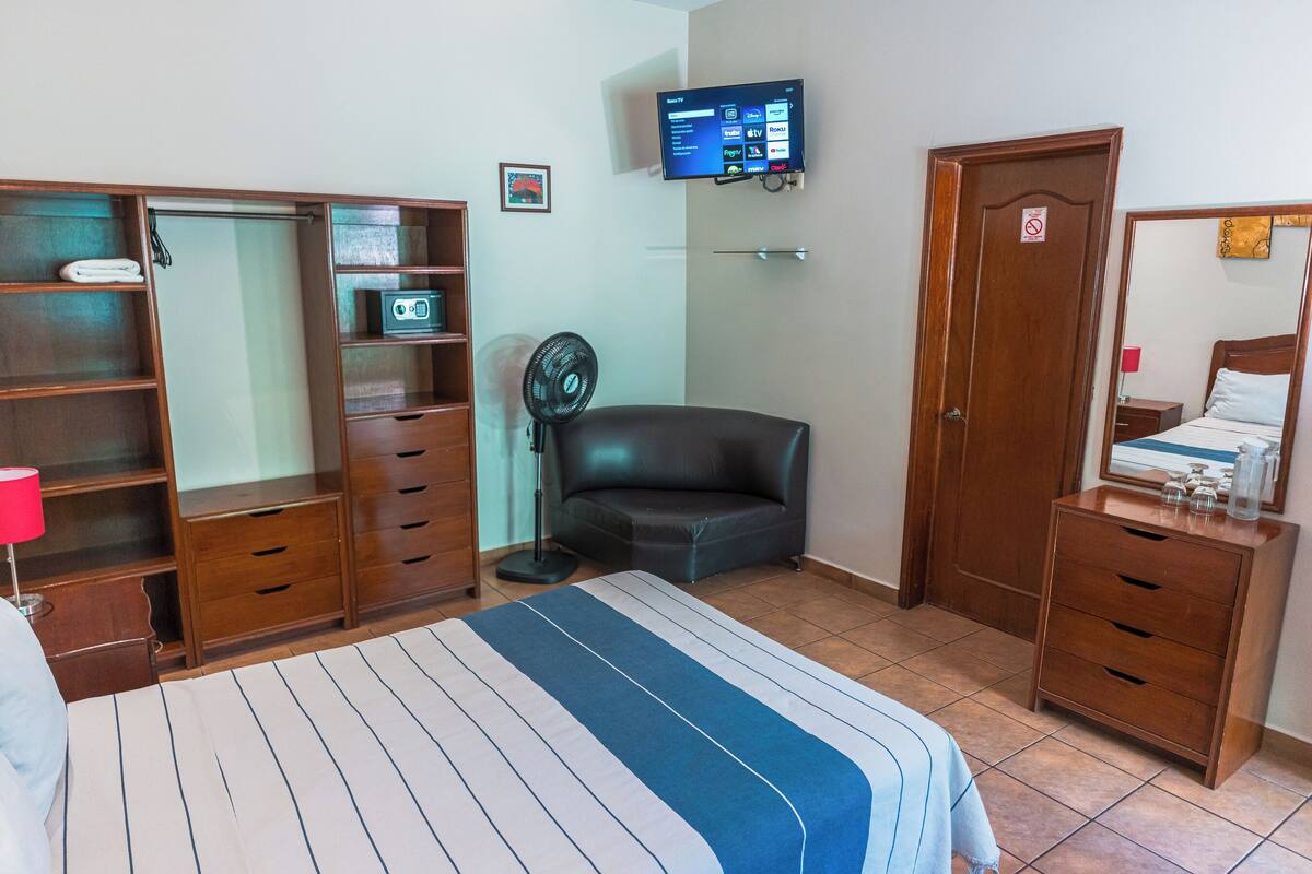 Habitación de hotel con cama, armario, escritorio, silla, televisor montado en la pared y puerta con un cartel.