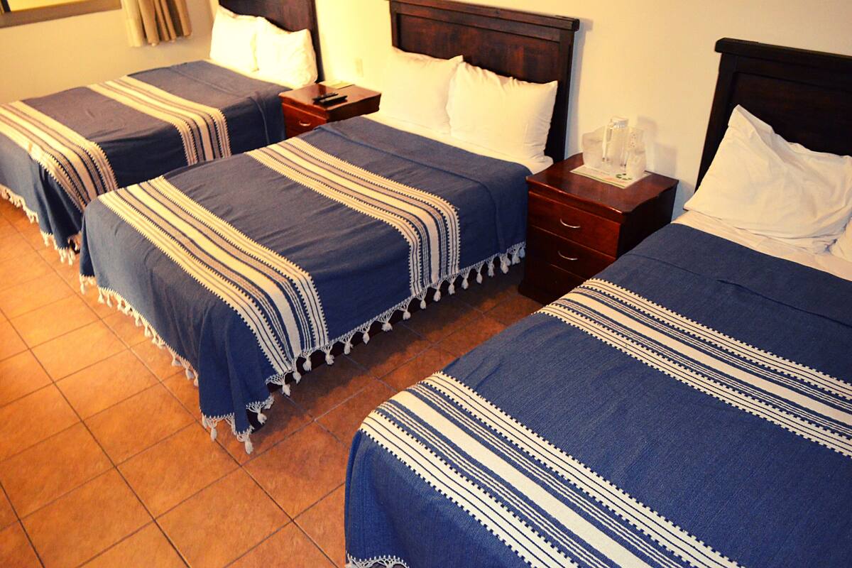 Una habitación de hotel con tres camas, cada una con una colcha azul y blanca a rayas. Hay mesitas de noche de madera con lámparas y una pequeña mesa con una jarrón y vasos.