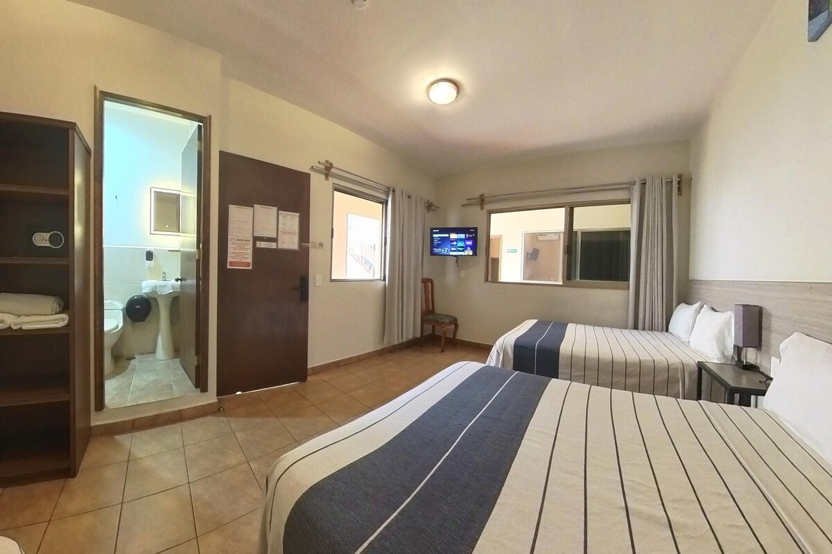 A hotel room with two beds, a TV, a desk, a wardrobe, and a bathroom visible through an open door.
