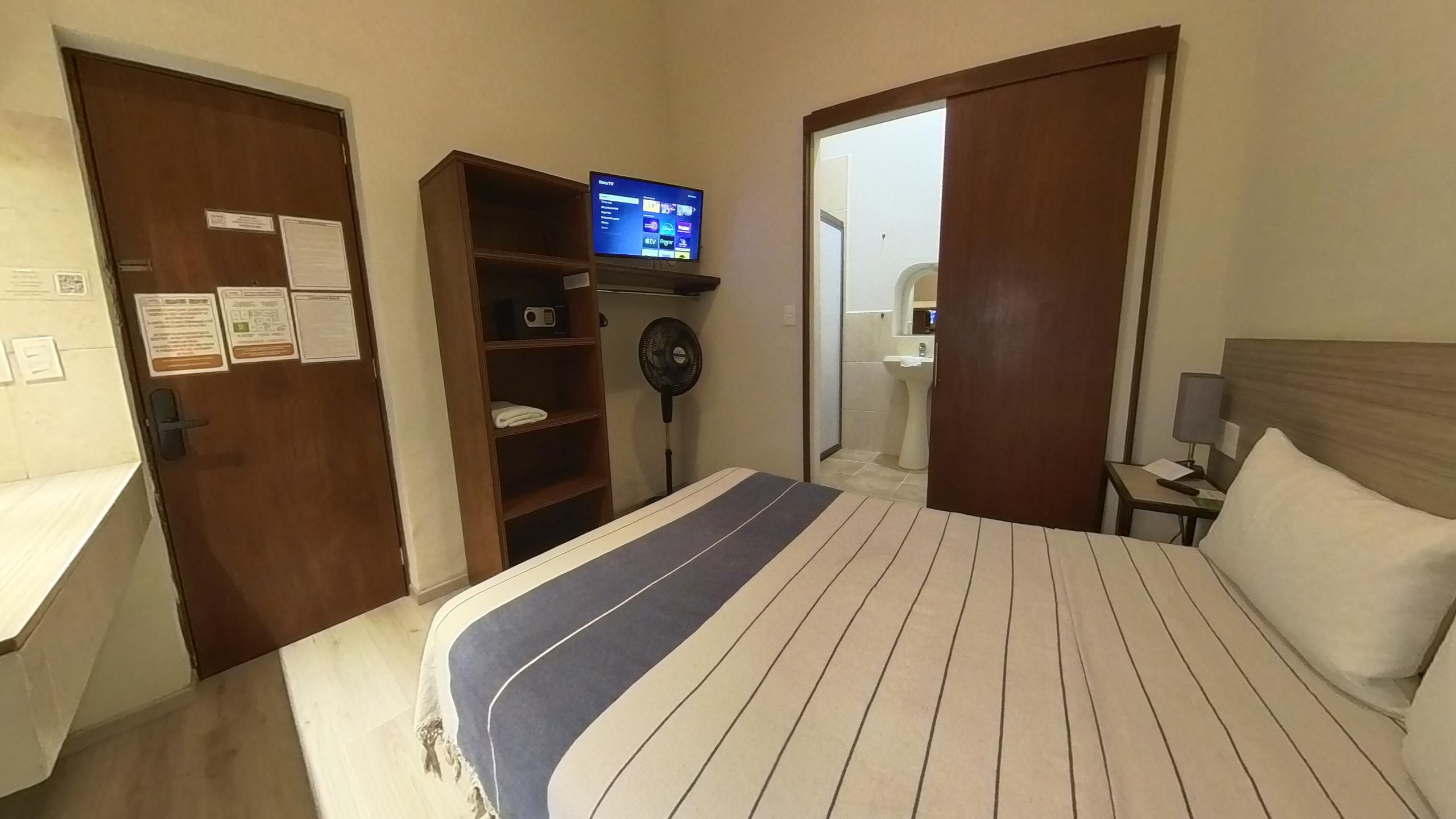Une chambre d’hôtel avec un lit, une télévision, un ventilateur, un bureau et une salle de bain visible à travers une porte ouverte.