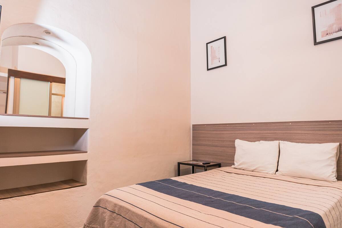 Una habitación de hotel con cama, una mesita, un espejo y un cuadro en la pared.