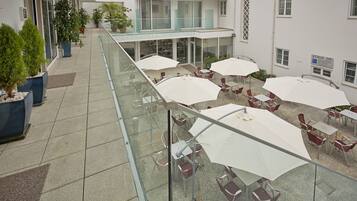 A glass-enclosed outdoor seating area with tables and chairs, potted plants, and a building with a sign reading "ZONA DE".