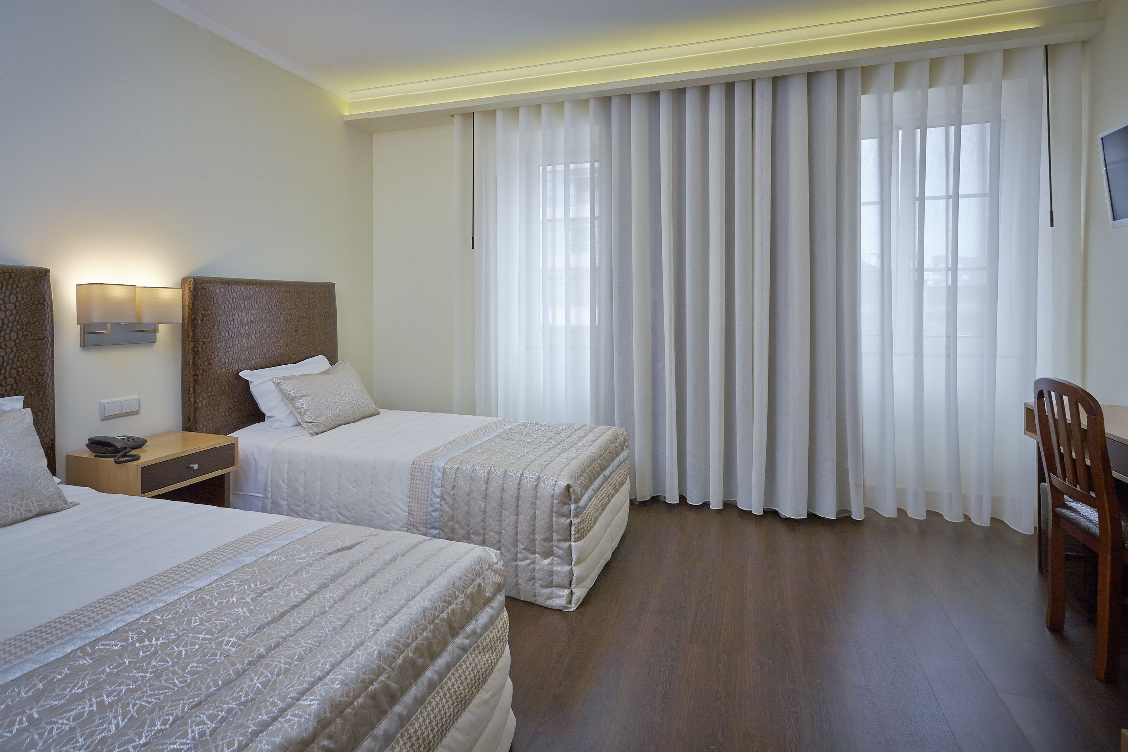 A hotel room with two beds, a desk, a chair, and a large window with curtains.