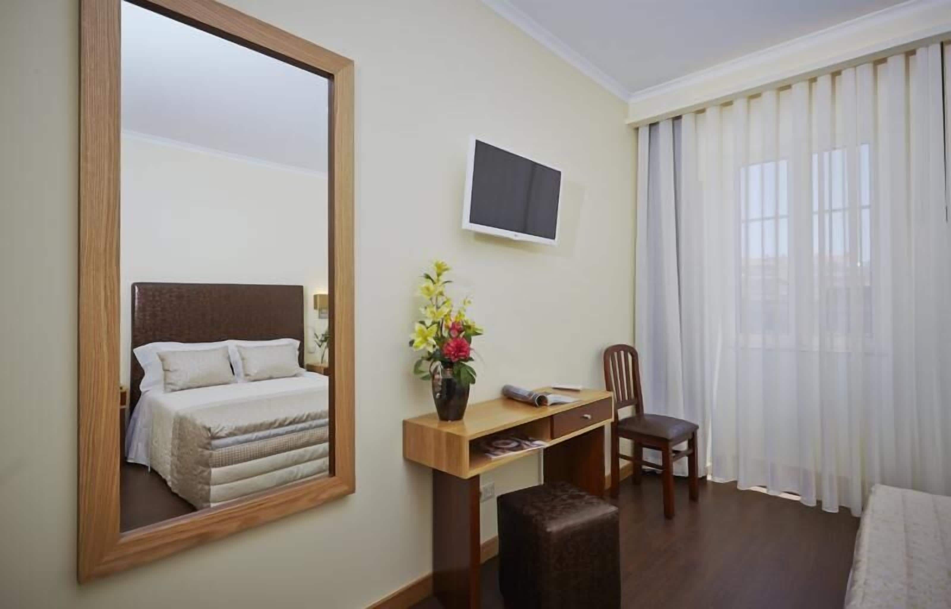 Habitación de hotel con cama, escritorio, silla, televisor y una ventana grande con cortinas.