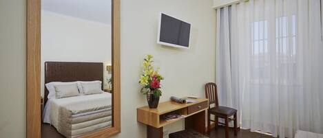 Habitación de hotel con cama, escritorio, silla, televisor y una ventana grande con cortinas.