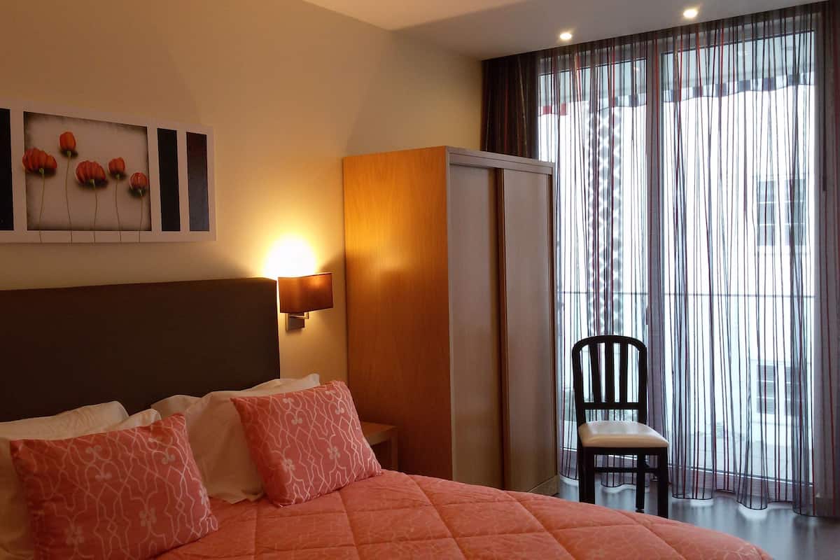 A hotel room with a bed, a chair, a painting, and a window with curtains.