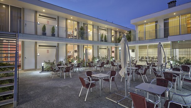 A modern outdoor cafe area with multiple tables and chairs, large windows, and potted plants.
