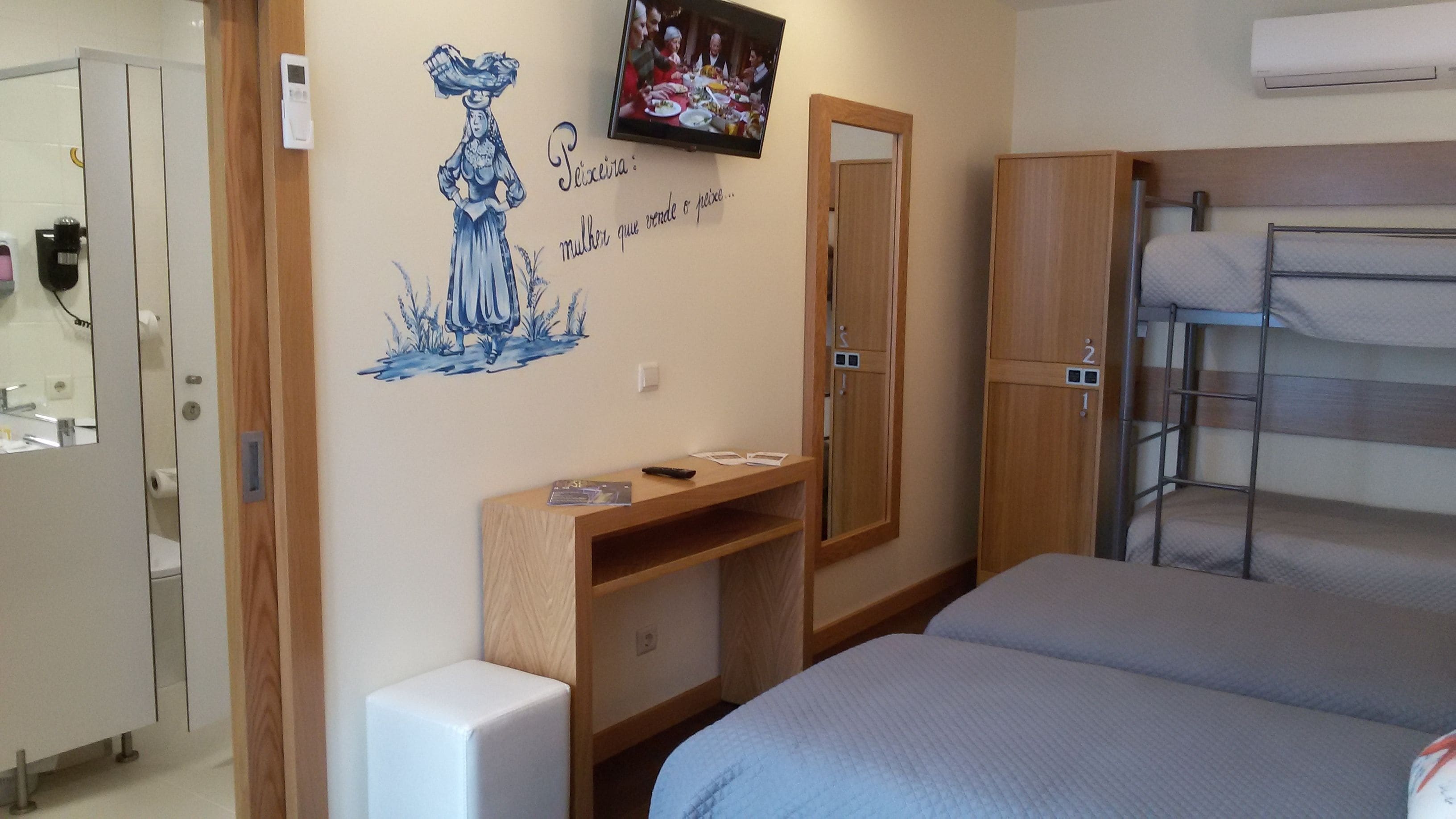 A hotel room with a bed, a TV mounted on the wall, a wooden desk, and a mirror.