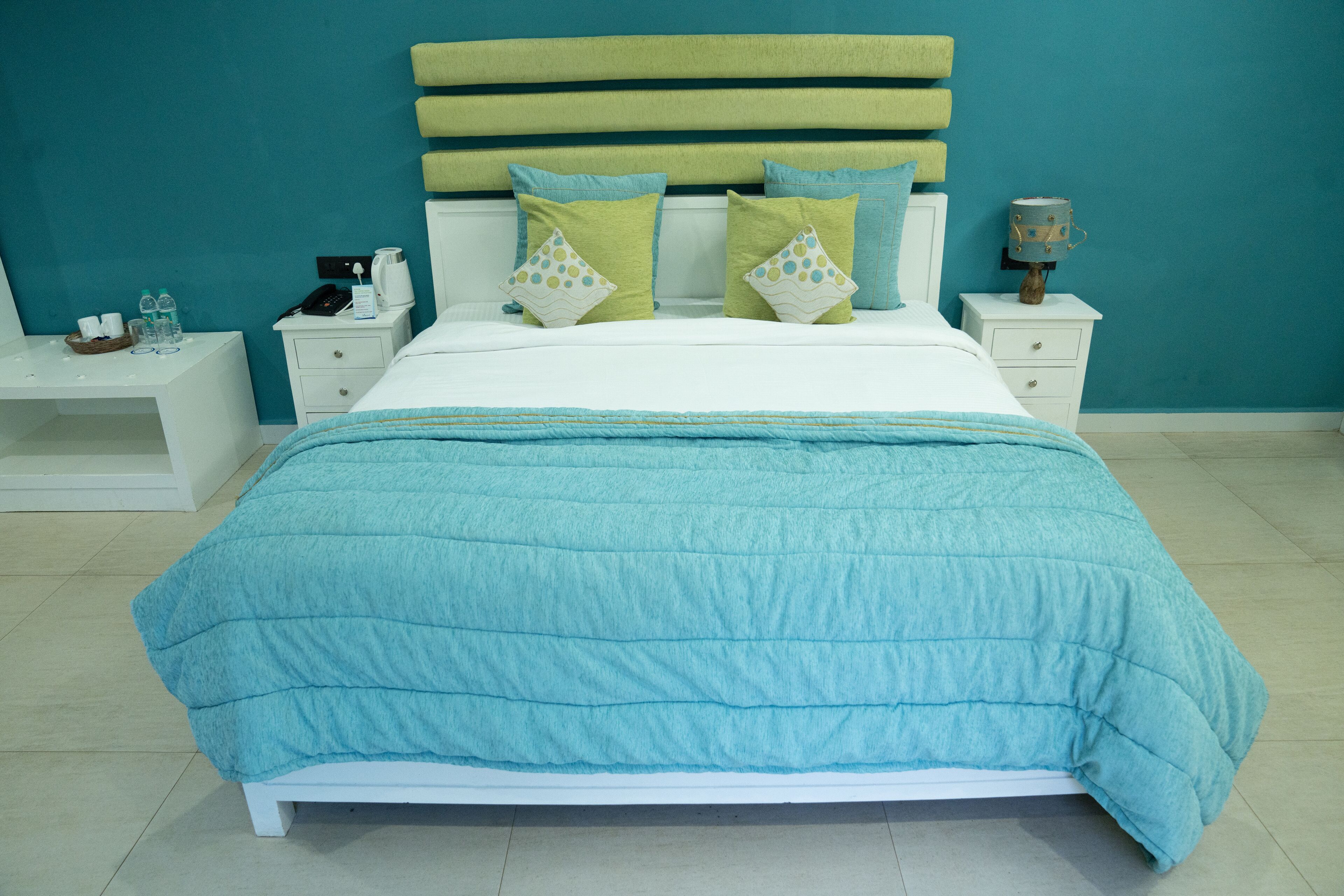 Una cama bien hecha con colcha color turquesa, cabecera blanca y dos almohadas a juego. Hay una mesita de noche con una lámpara y un pequeño atril con un teléfono y una taza.