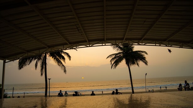 A beach scene with a covered area, palm trees, and people relaxing. - Regenta Beach House Resort, Goa (Colva)