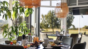 Una mesa de comedor dispuesta con platos, vasos y una jarrón, con vistas a follaje y un edificio.