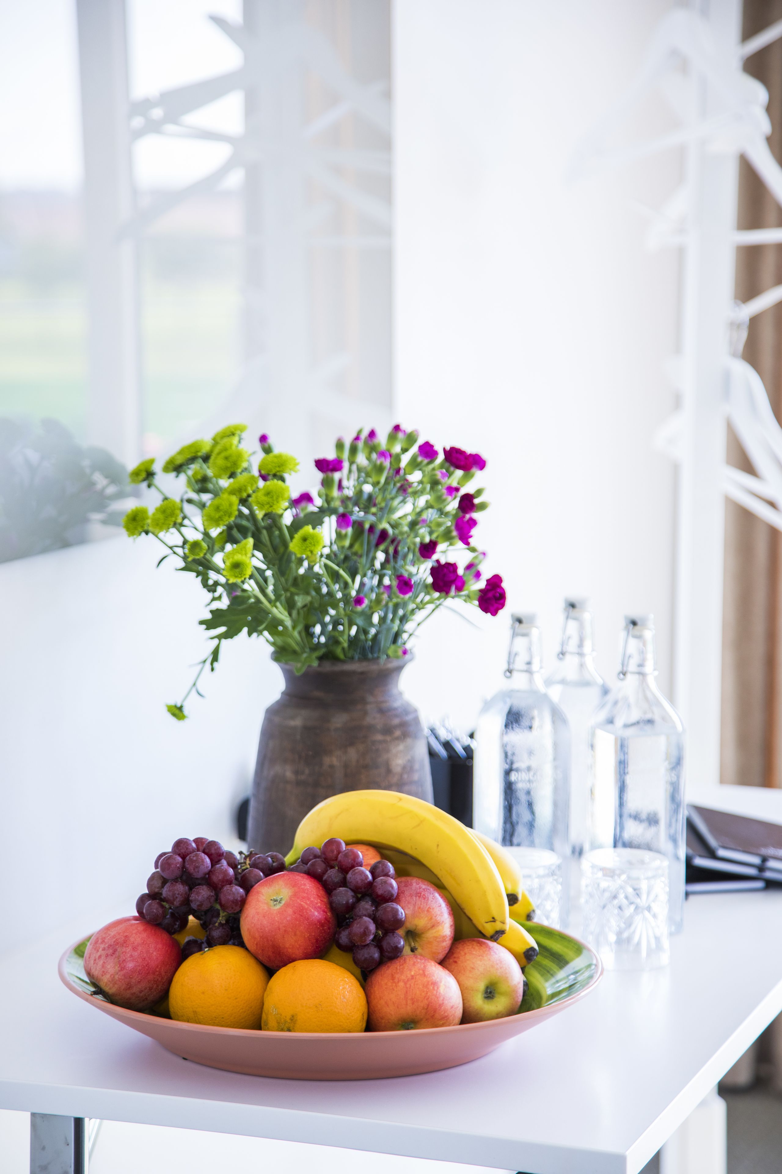A table with a fruit bowl, a vase with flowers, and clear glass bottles.