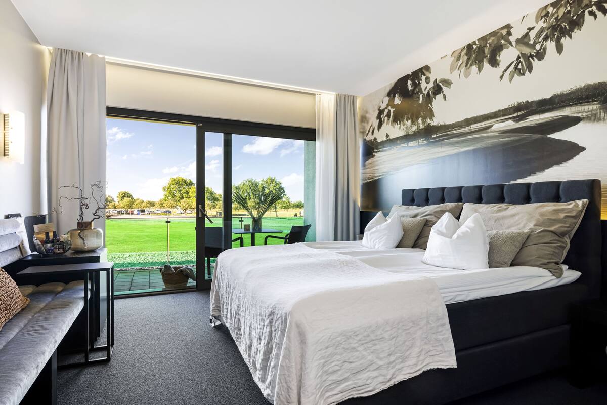 A hotel room with a large bed, a sofa, a small table with a vase, and a view of a green landscape through a glass door.
