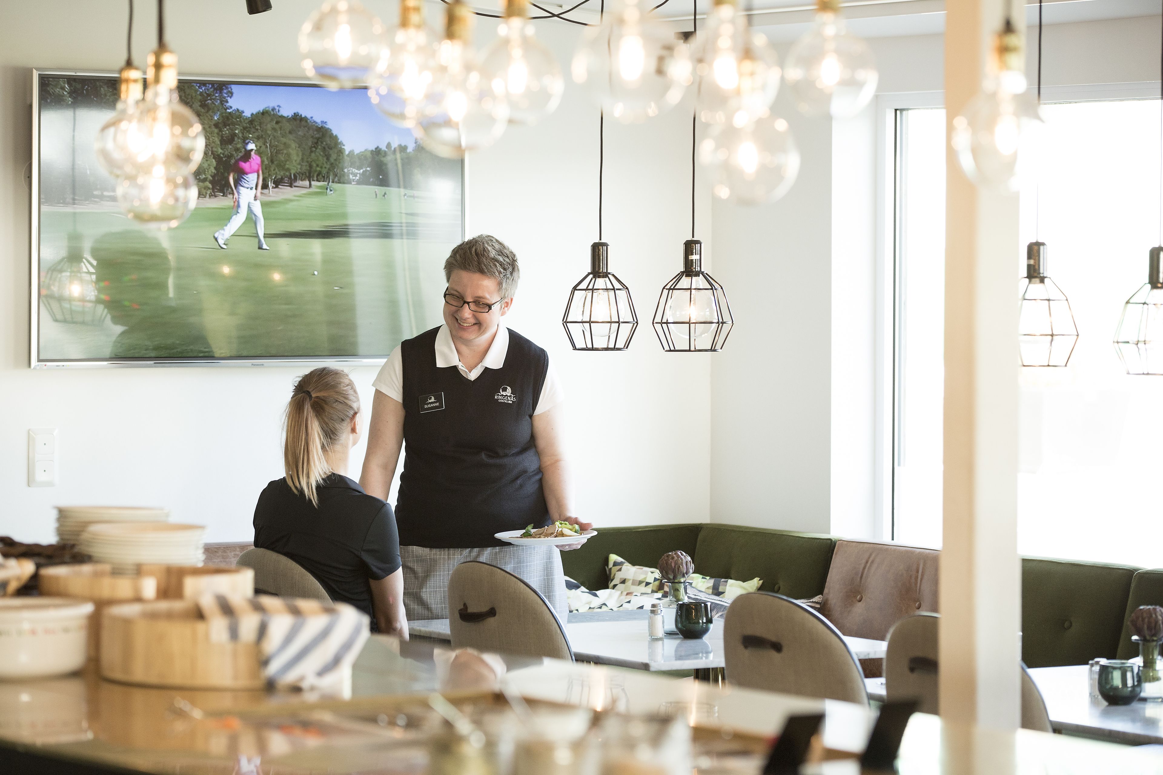 A modern dining area with a large TV displaying a golf game, pendant lighting, and a casual seating arrangement.