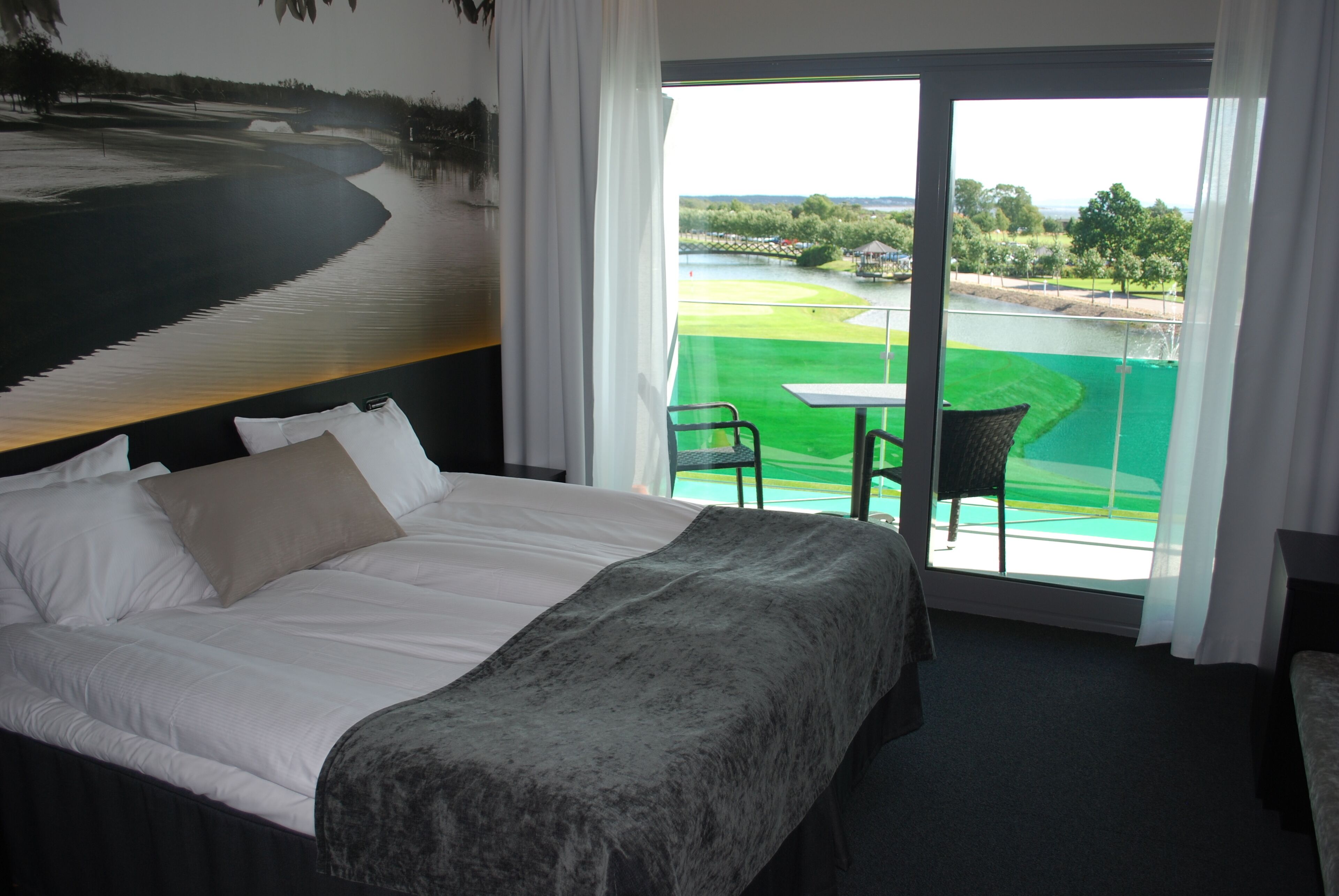 Ein Hotelzimmer mit einem Bett, einem Sessel und einem Tisch. Durch ein großes Fenster bietet das Zimmer Blick auf einen Golfplatz.
