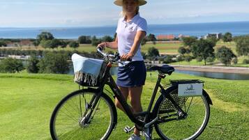 A person standing next to a bicycle with a basket, on a grassy area with a view of the sea and buildings in the background.