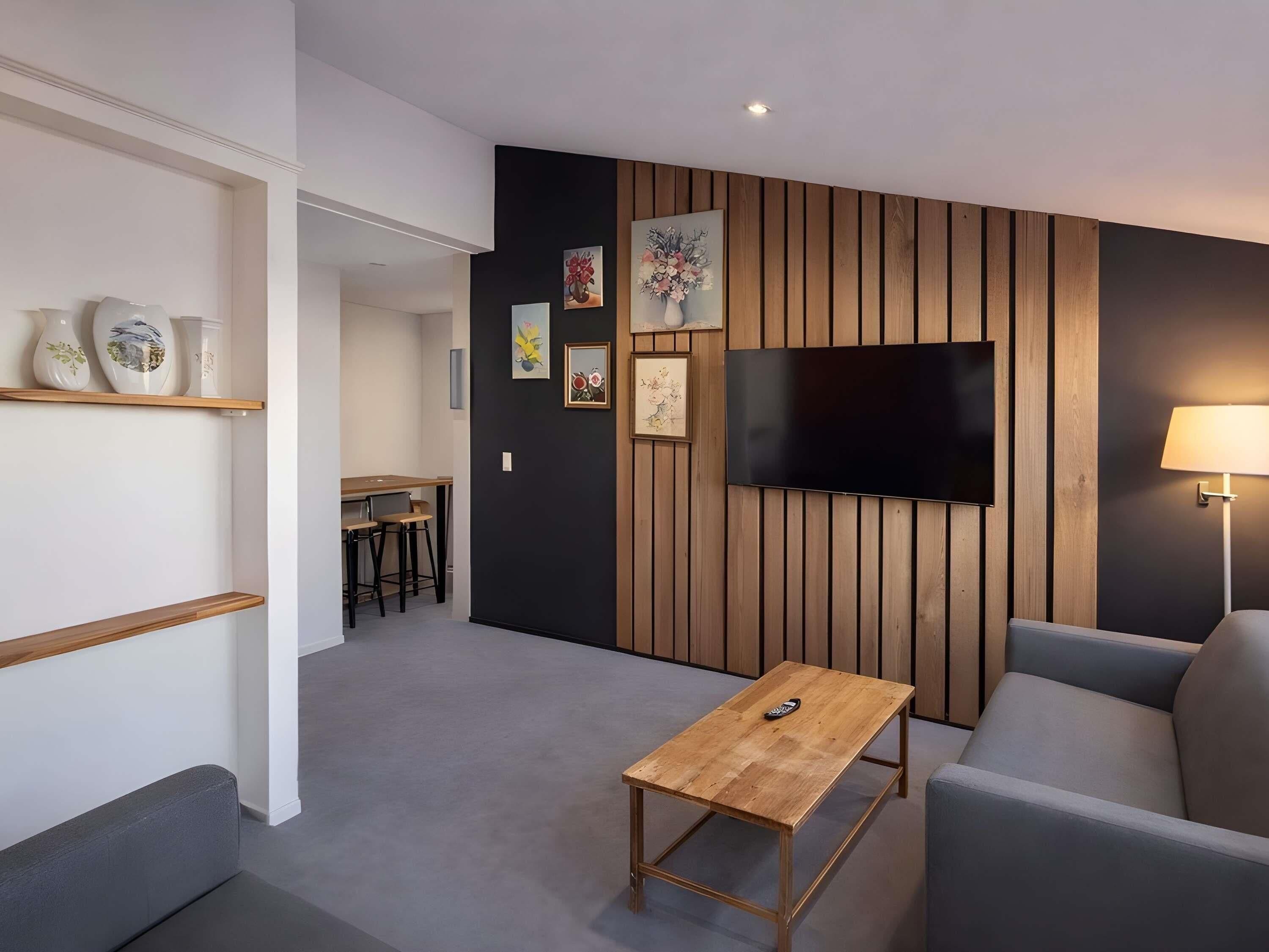 Una sala de estar moderna con un televisor de pantalla plana, una mesa de centro de madera, un sofá gris y un comedor al fondo.