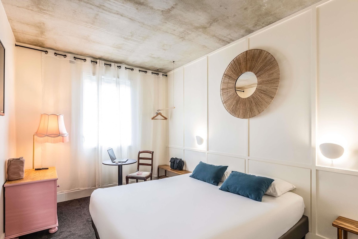 A hotel room with a large bed, a desk with a laptop, a chair, a small table, and a hanging light fixture.